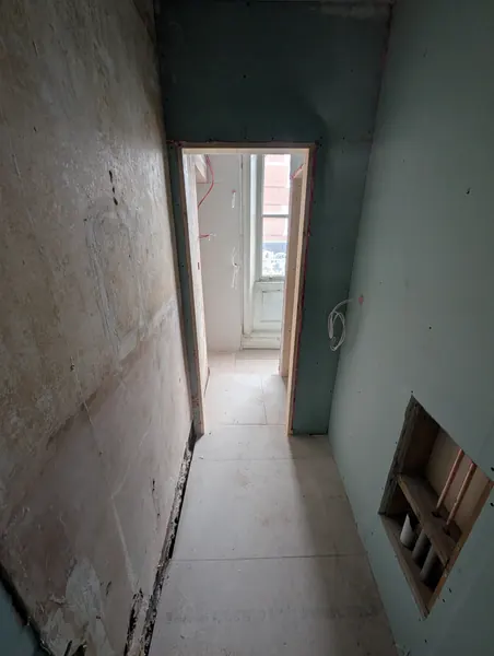 A corridor between a stripped wall and new boards, with an opening showing pipework and a doorway to a window beyond