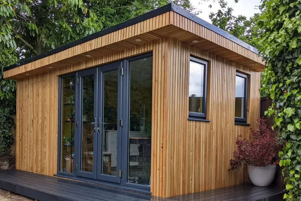 Cedar-clad garden room with grey bifold doors, a black composite deck and an ivy hedge behind