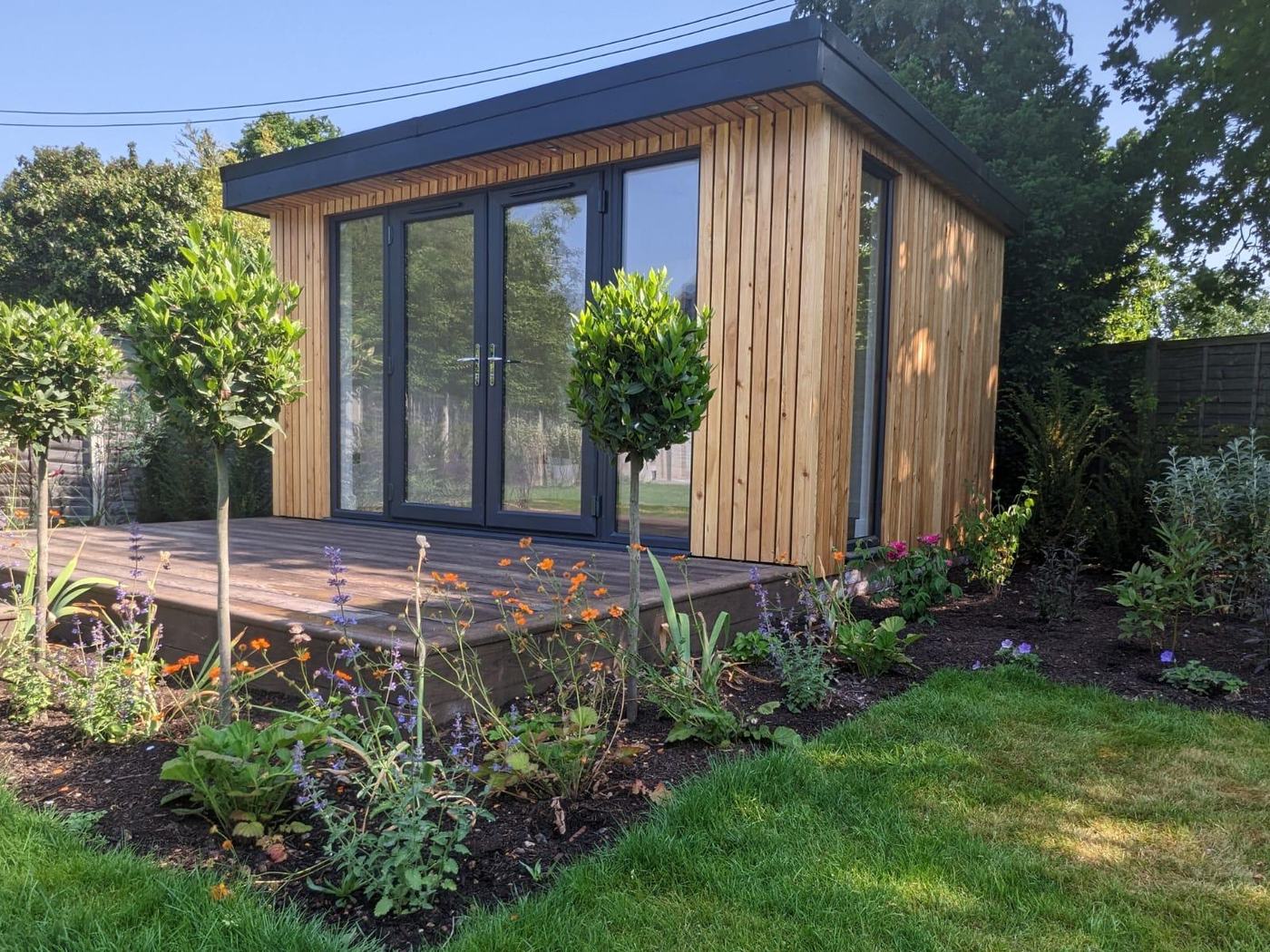 A finished garden room with timber cladding and a flat roof, behind a planted border