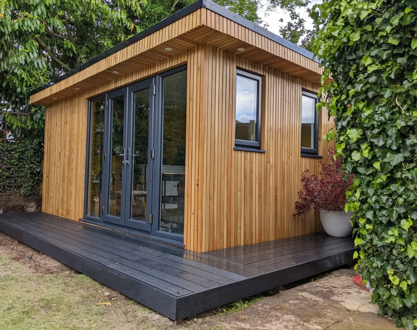 Finished cedar-clad garden room with a black composite deck, framed by an ivy hedge