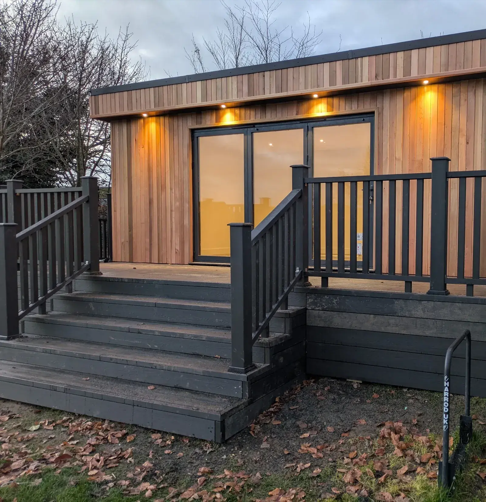A cedar-clad bespoke garden room at dusk, warm light glowing through full-height glazing onto a dark composite deck